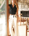 Woman in black sleeveless top and white pants standing in a room with stone wall and wooden furniture.