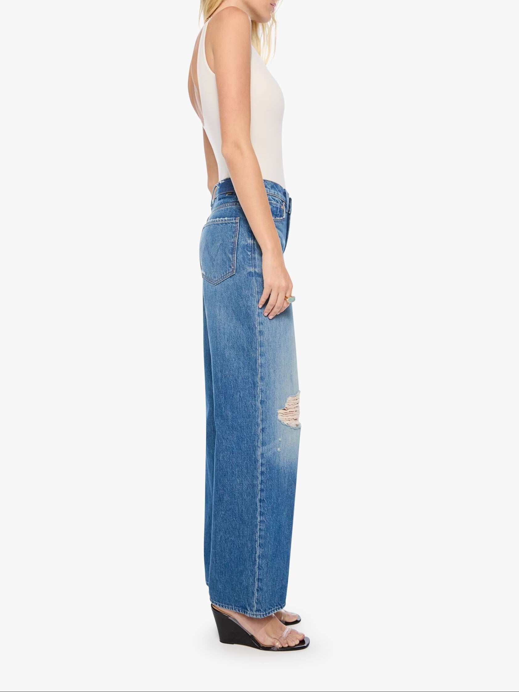 A woman shown in profile wearing a white sleeveless top and high-waisted, wide-leg blue denim jeans with a distressed knee detail, set against a plain light gray background.