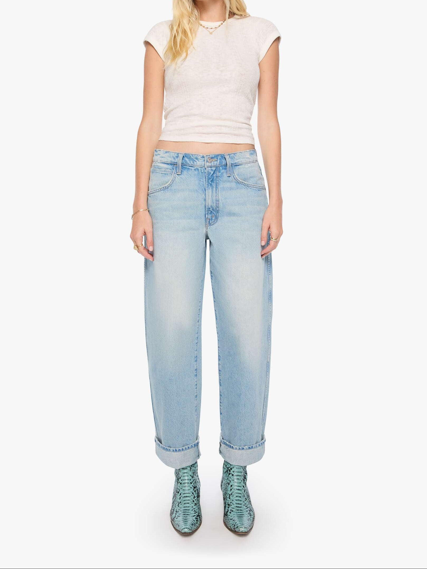 A woman modeling light-wash blue jeans with a cream ribbed crop top and green snakeskin boots, standing against a plain white studio background.