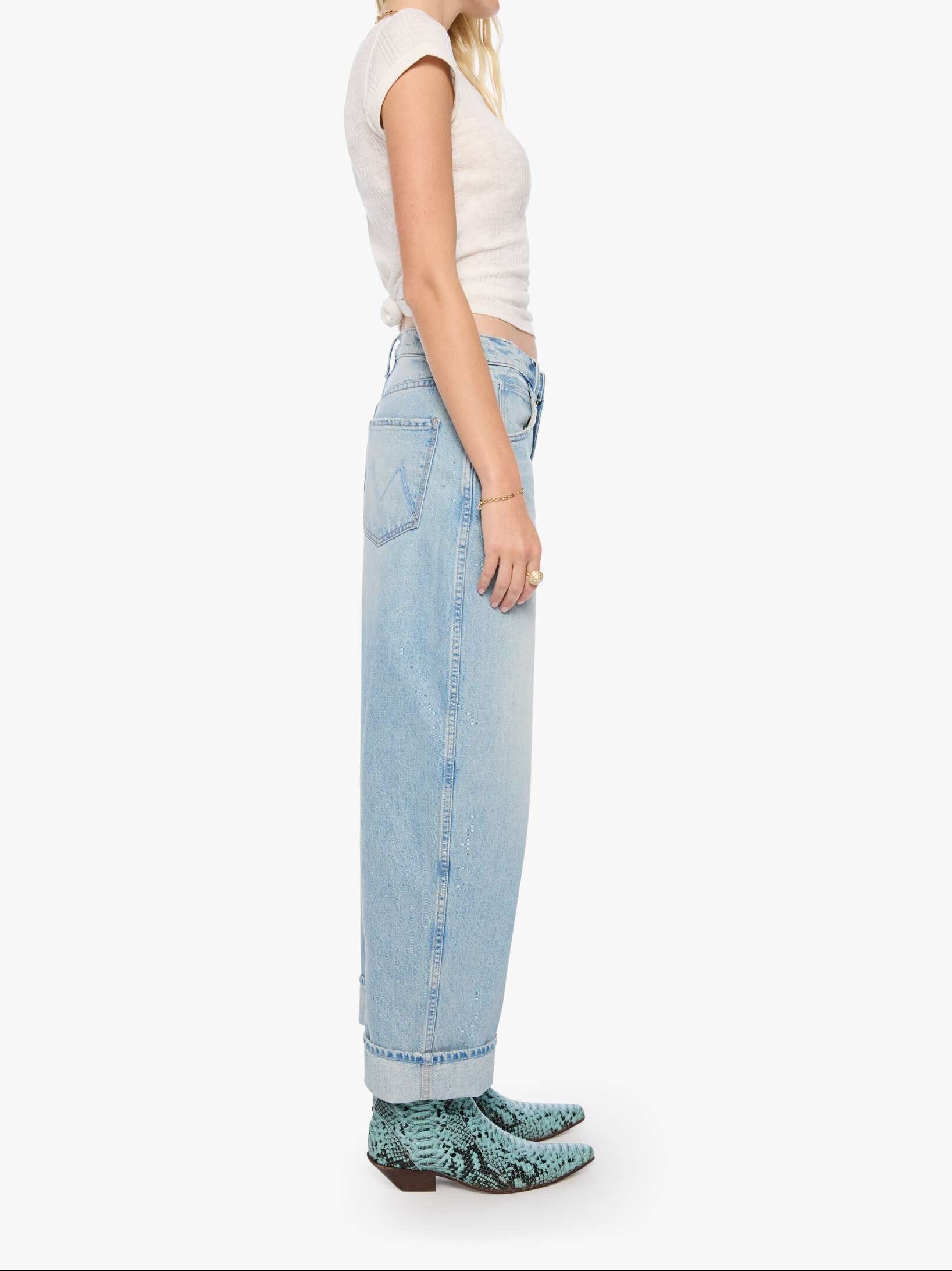 A woman shown in profile wearing a white sleeveless top, light-wash wide-leg jeans, and green snakeskin-patterned shoes against a plain light gray studio background.