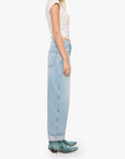 A woman shown in profile wearing a white sleeveless top, light-wash wide-leg jeans, and green snakeskin-patterned shoes against a plain light gray studio background.