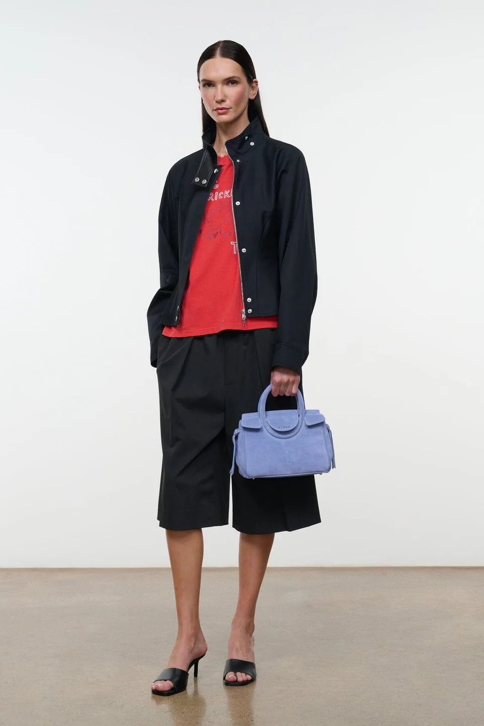 A model stands in a studio wearing a black cropped jacket over a red graphic T-shirt, black shorts, and black heeled sandals, while holding a light blue structured handbag.