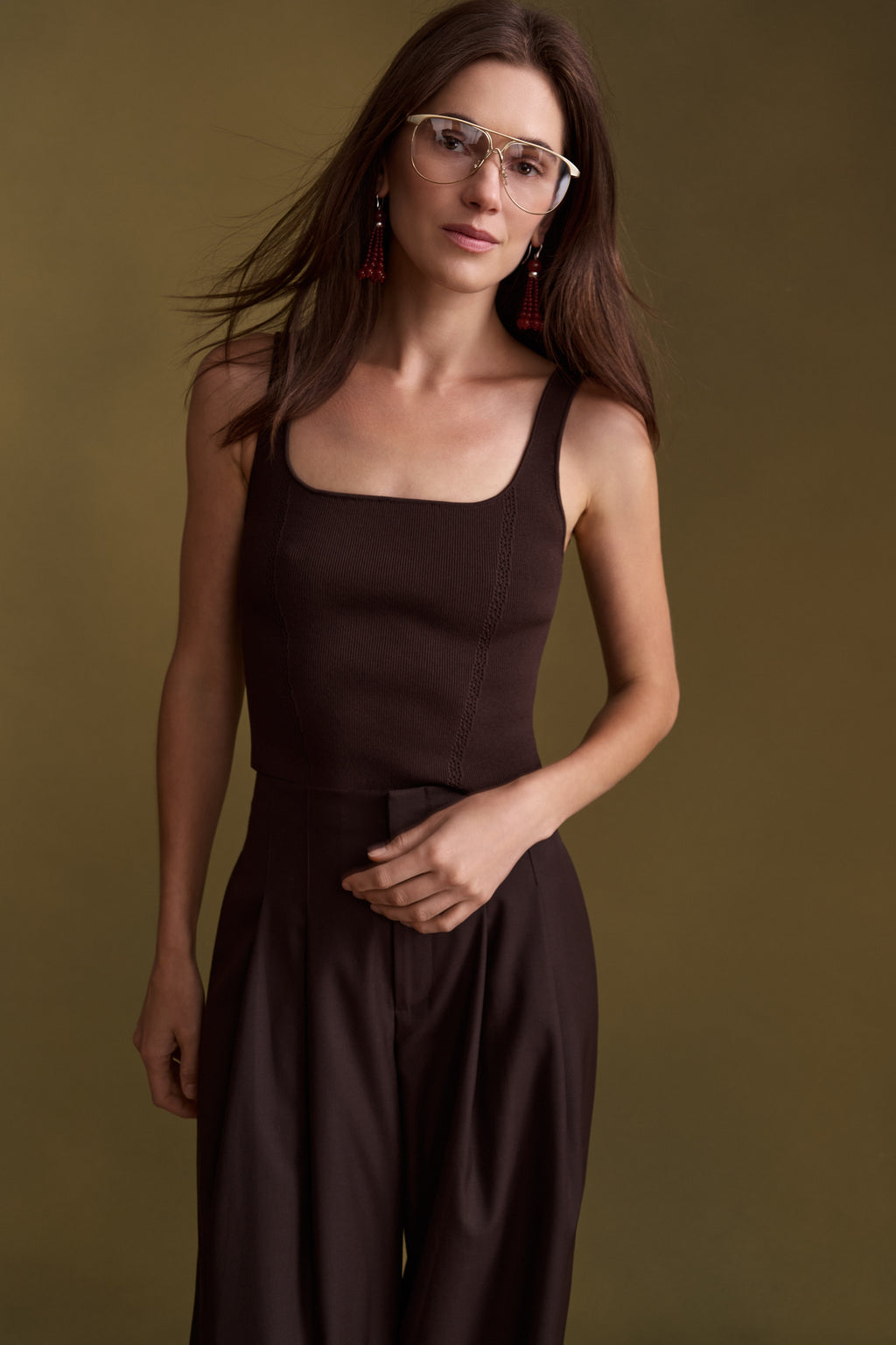 A woman wearing a dark brown sleeveless jumpsuit with a square neckline and wide legs, paired with clear-framed glasses and red tassel earrings, posed against a solid olive green background.