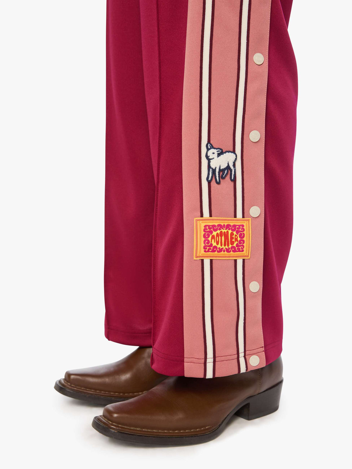 Close-up of a person's legs wearing pink and burgundy striped pants with decorative patches and brown leather boots, photographed against a clean white background.