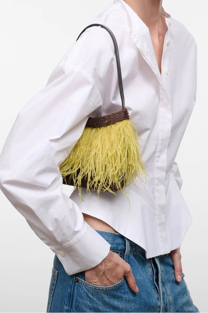 A model wearing a white button-down shirt and blue jeans, carrying a small yellow fringe handbag with a dark brown top band and black strap against a plain white studio background.