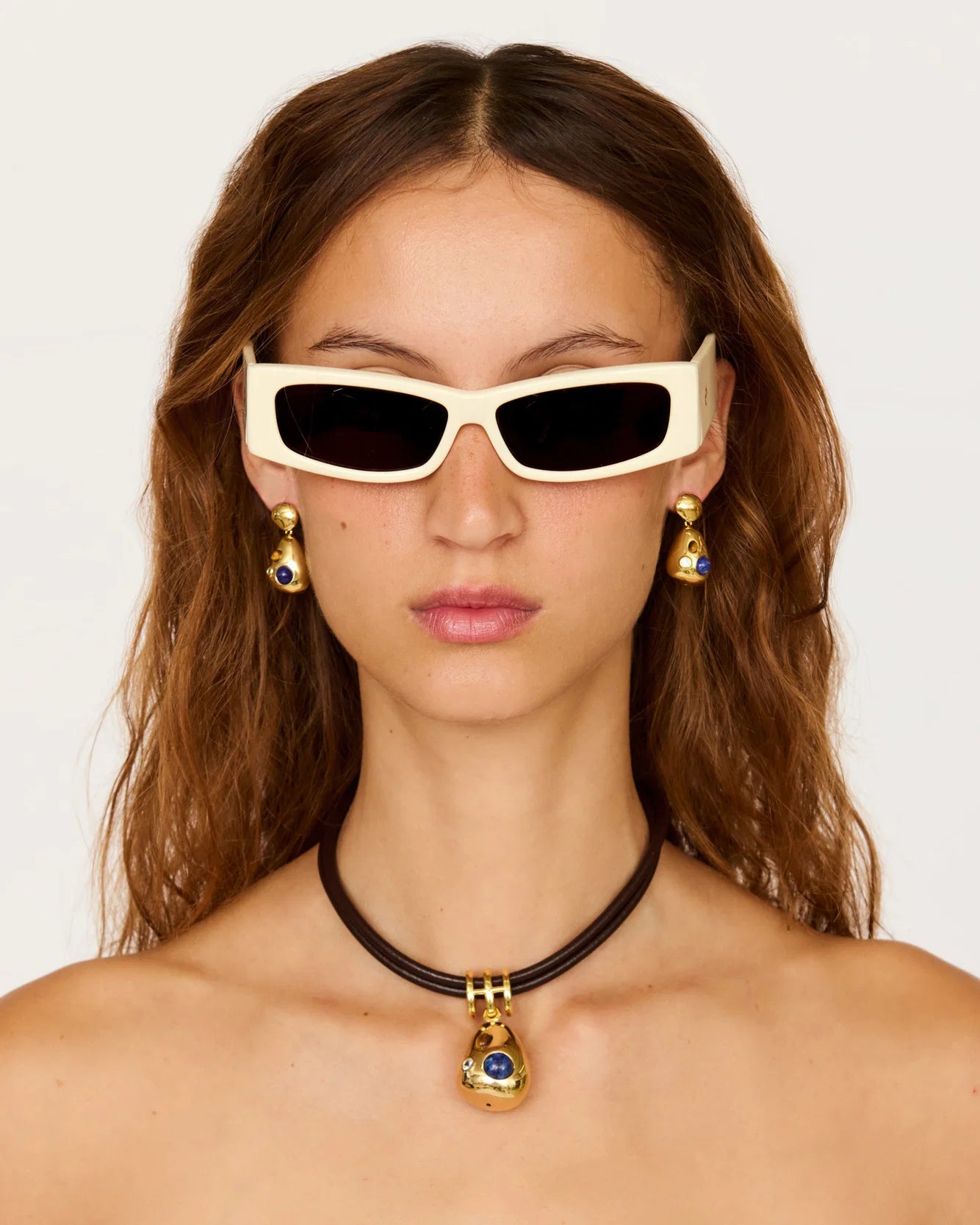 A close-up of a model wearing white cat-eye sunglasses with black lenses, gold drop earrings, and a black choker necklace with a gold pendant featuring a blue gemstone, against a plain light background.