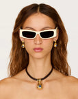 A close-up of a model wearing white cat-eye sunglasses with black lenses, gold drop earrings, and a black choker necklace with a gold pendant featuring a blue gemstone, against a plain light background.