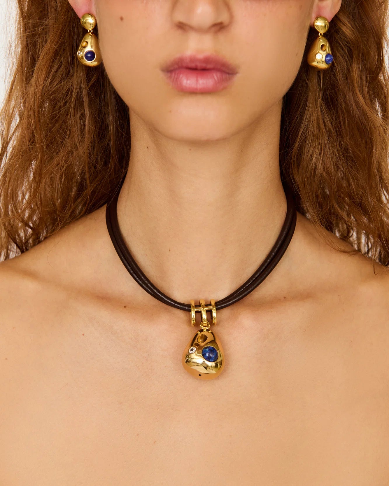 A close-up of a model wearing a black cord choker necklace with a gold teardrop pendant featuring a blue stone, along with matching gold and blue earrings.