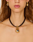 A close-up of a model wearing a black cord choker necklace with a gold teardrop pendant featuring a blue stone, along with matching gold and blue earrings.