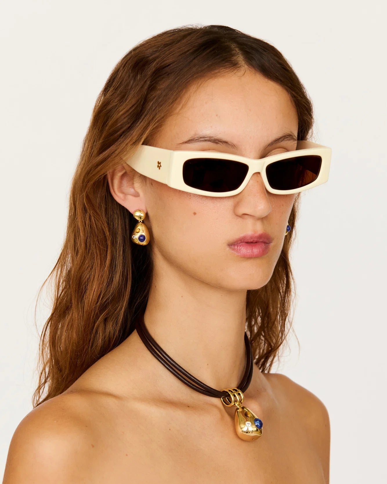 A close-up of a model wearing cream-colored cat-eye sunglasses with dark lenses, gold earrings, and a black cord necklace with a gold and blue pendant, set against a plain light background.