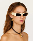 A close-up of a model wearing cream-colored cat-eye sunglasses with dark lenses, gold earrings, and a black cord necklace with a gold and blue pendant, set against a plain light background.