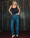 A woman wearing a black tank top, blue straight-leg jeans, and black pointed-toe flats, standing in front of a dark metal gate.