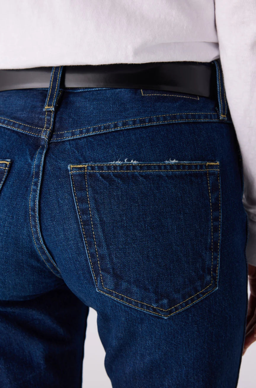 Close-up back view of dark blue denim jeans worn with a black belt and white top, showing the back pocket and stitching details.