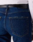 Close-up back view of dark blue denim jeans worn with a black belt and white top, showing the back pocket and stitching details.