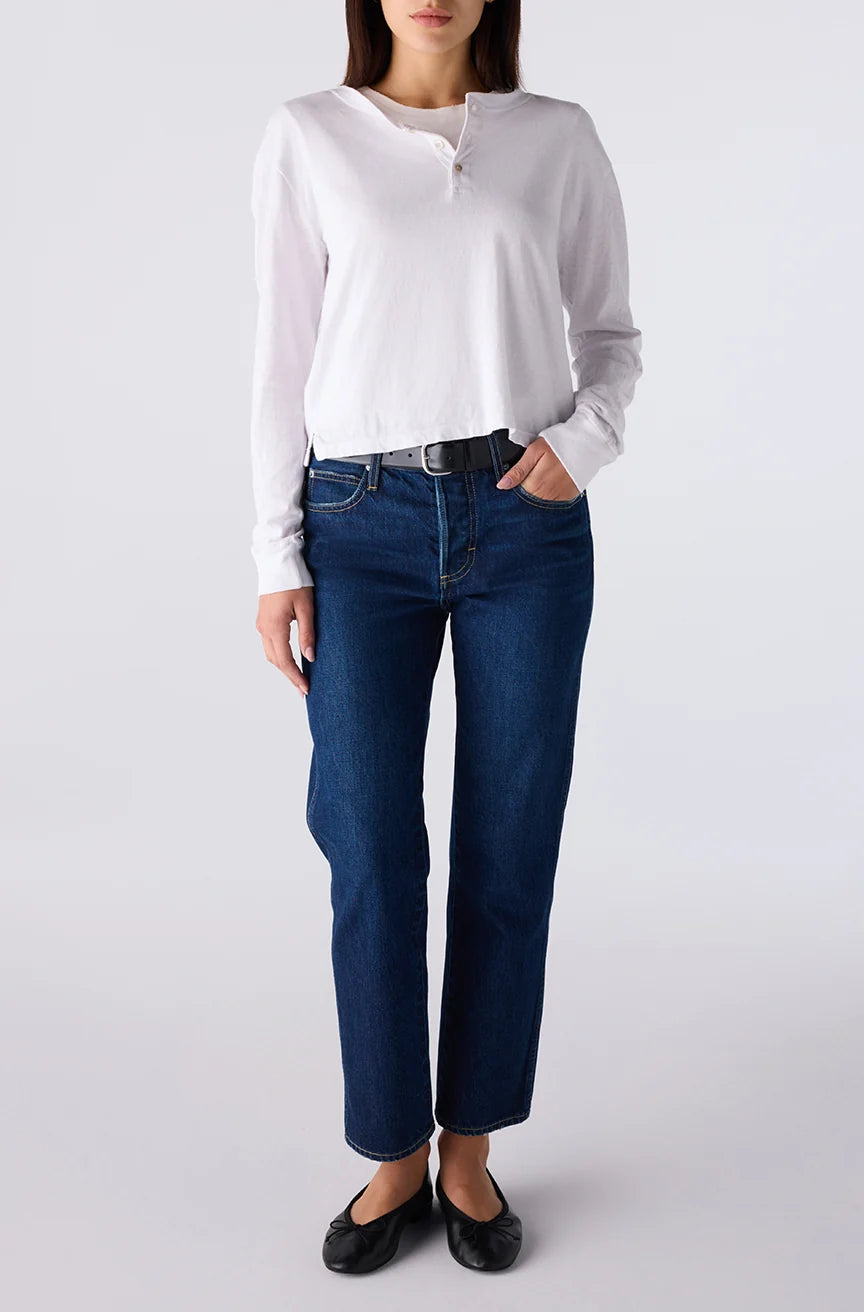 A model wearing a white long-sleeve henley shirt, dark blue straight-leg jeans, a black belt, and black flats, standing against a light gray studio background.