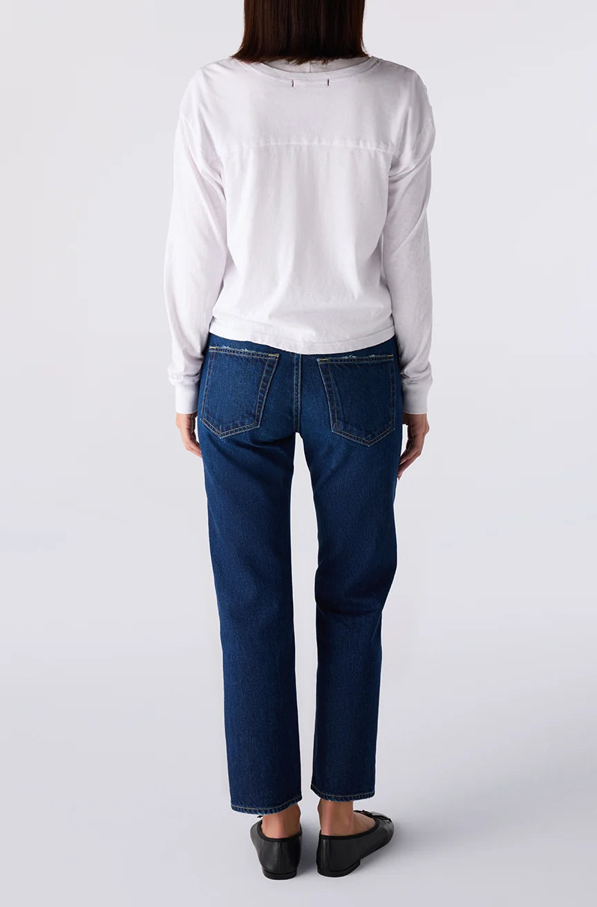 A person shown from the back wearing a white long-sleeve top, dark blue straight-leg jeans, and black flats, photographed against a light gray studio background.