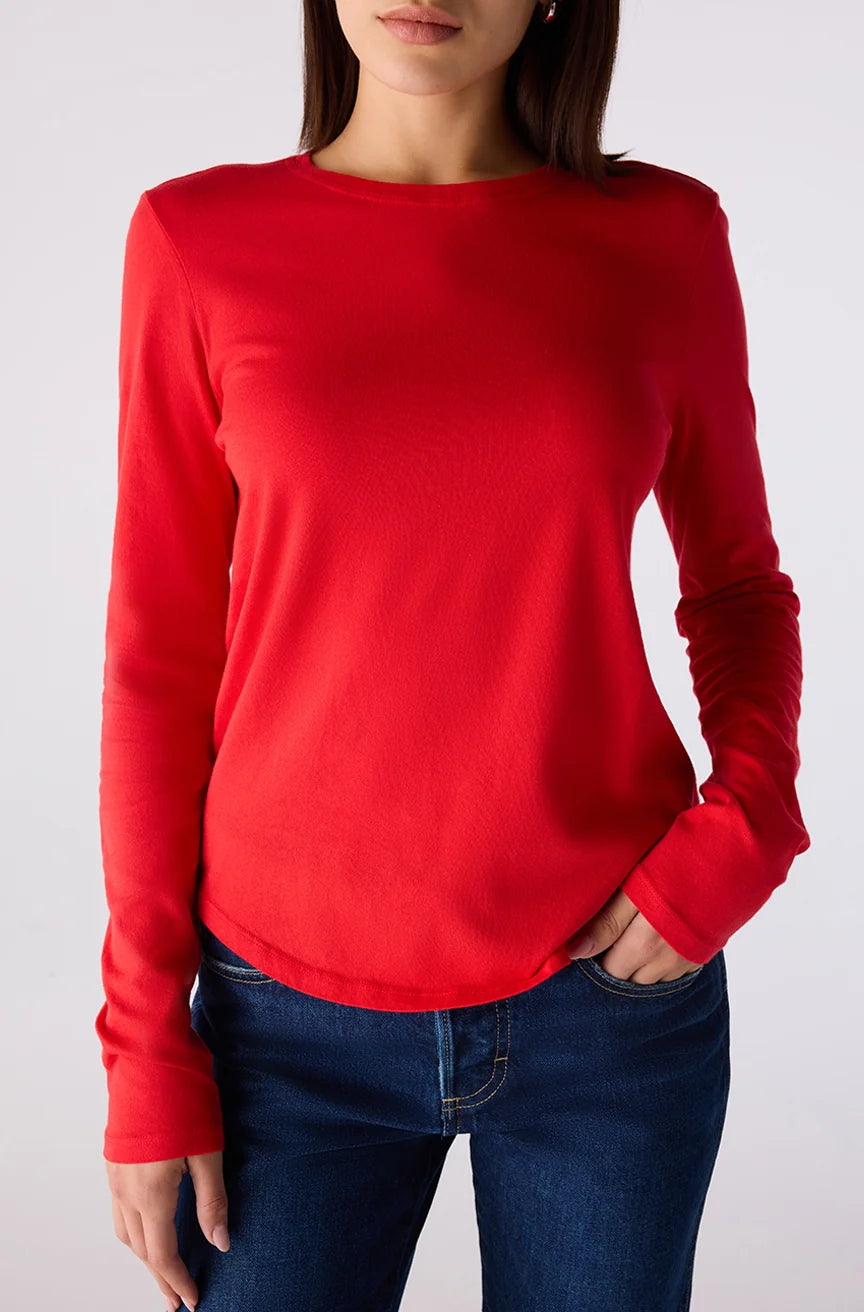 A woman wearing a bright red long-sleeve crewneck top and dark blue jeans, shown from the neck down against a light gray studio background.