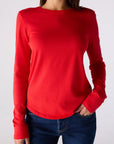 A woman wearing a bright red long-sleeve crewneck top and dark blue jeans, shown from the neck down against a light gray studio background.