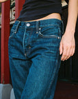 A close-up of a person wearing dark blue denim jeans with a black top, standing in front of a red and black background.