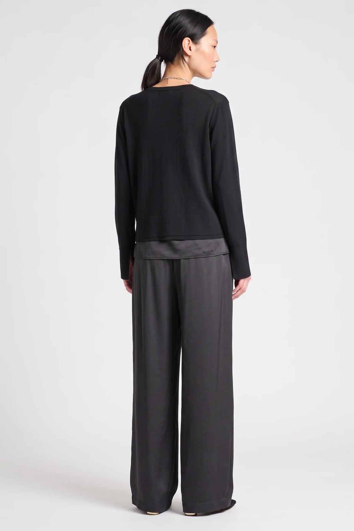 model wearing a black vneck sweater over a silky black top with black pants on a grey background, back view