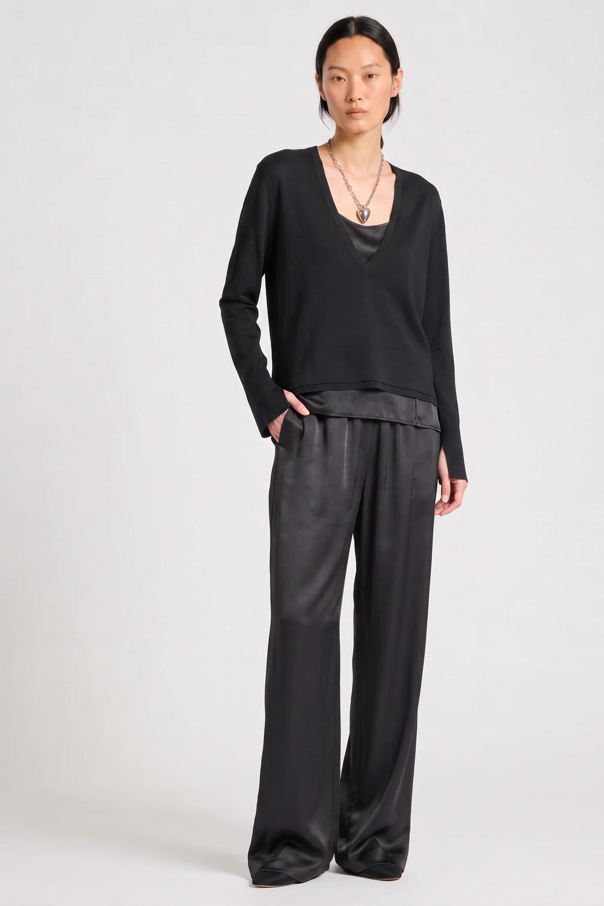 model wearing a black vneck sweater over a silky black top with black pants on a grey background
