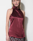 A woman modeling a sleeveless, high-neck satin top in a deep burgundy color, paired with a brown and white plaid skirt, against a plain off-white background.