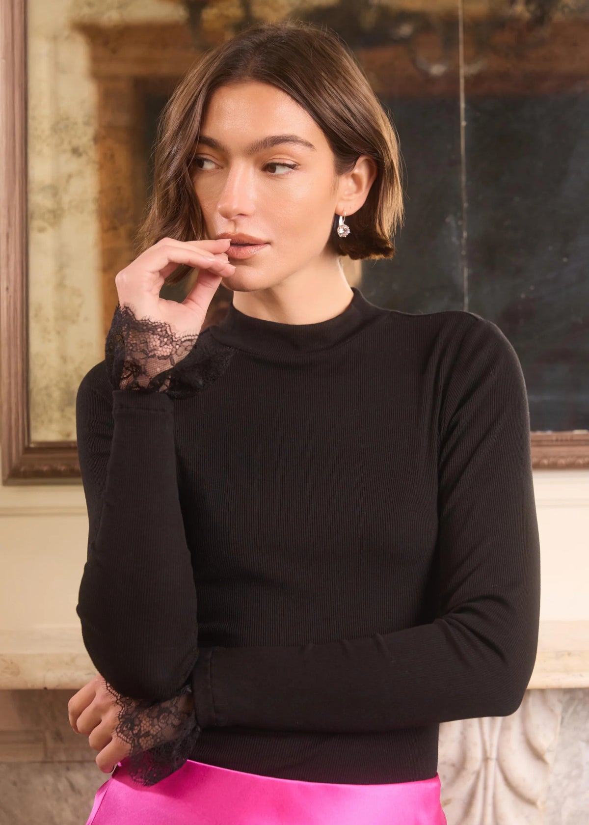 A woman wearing a black mock-neck long-sleeve top with black lace ruffle cuffs, paired with a bright pink skirt, posed in front of an indoor mirror.