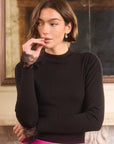 A woman wearing a black mock-neck long-sleeve top with black lace ruffle cuffs, paired with a bright pink skirt, posed in front of an indoor mirror.