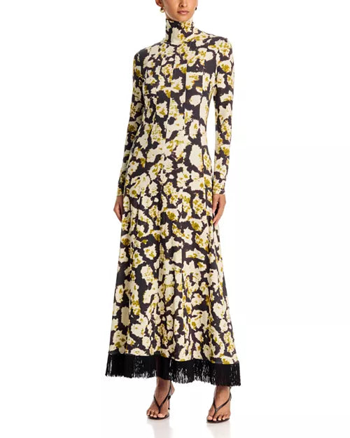 A long-sleeve high-neck maxi dress with a black, cream, and olive yellow floral pattern and a black fringe hem, modeled on a white background.