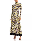 A long-sleeve high-neck maxi dress with a black, cream, and olive yellow floral pattern and a black fringe hem, modeled on a white background.