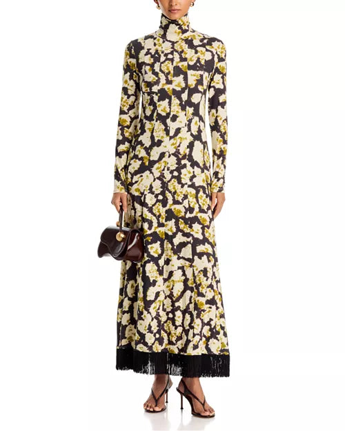A model wears a long-sleeved high-neck maxi dress featuring a black base with a yellow and cream floral print and a black fringe hem. She is holding a small dark brown handbag and wearing black strappy heels against a plain white background.
