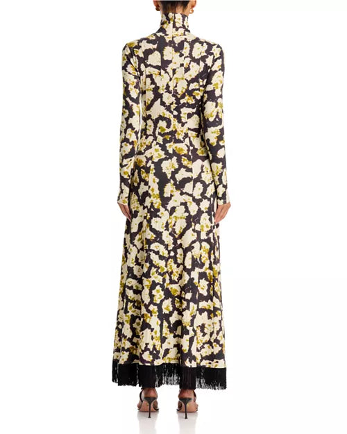 A long-sleeved maxi dress with a high mock neck, featuring an all-over floral print in cream and olive green on a black background, with black fringe detailing along the hem. The dress is shown from the back on a model against a white studio background.