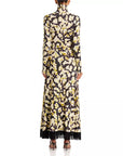 A long-sleeved maxi dress with a high mock neck, featuring an all-over floral print in cream and olive green on a black background, with black fringe detailing along the hem. The dress is shown from the back on a model against a white studio background.