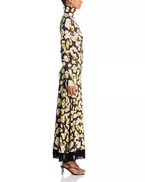 A long-sleeved maxi dress with a high mock neck, featuring an all-over floral print in cream and olive green on a black background, with black fringe detailing along the hem. The dress is shown from the back on a model against a white studio background, side view