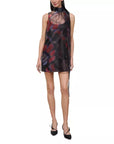 A model wearing a sleeveless high-neck mini dress made of sheer black mesh with a red and blue abstract floral print, paired with black lace-up heels, against a plain white background.