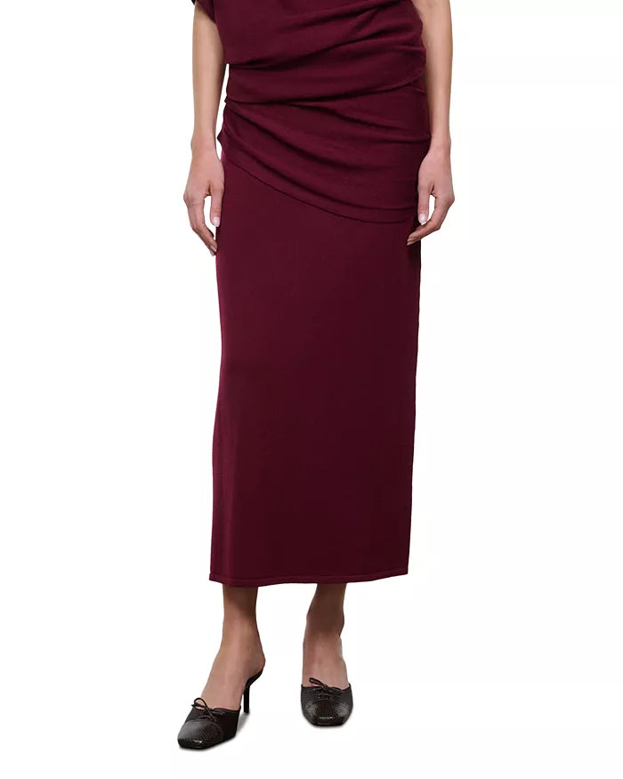 A woman wearing a draped midi skirt in deep burgundy, paired with black pointed-toe flats, shown against a plain white background.