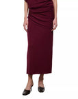 A woman wearing a draped midi skirt in deep burgundy, paired with black pointed-toe flats, shown against a plain white background.