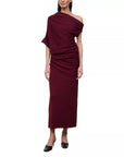 A woman modeling a draped one-shoulder midi skirt and top in deep burgundy, paired with black pointed-toe flats, against a plain white background.