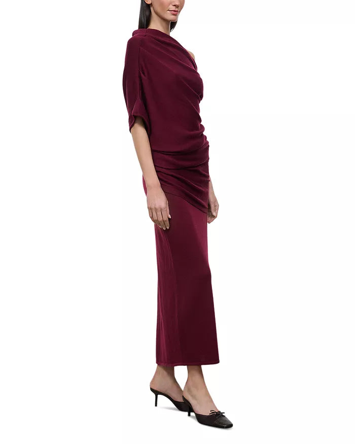 A woman modeling a draped, long-sleeved maxi dress in deep burgundy velvet, paired with black pointed-toe heels, against a plain white background.