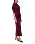 A woman modeling a draped, long-sleeved maxi dress in deep burgundy velvet, paired with black pointed-toe heels, against a plain white background.