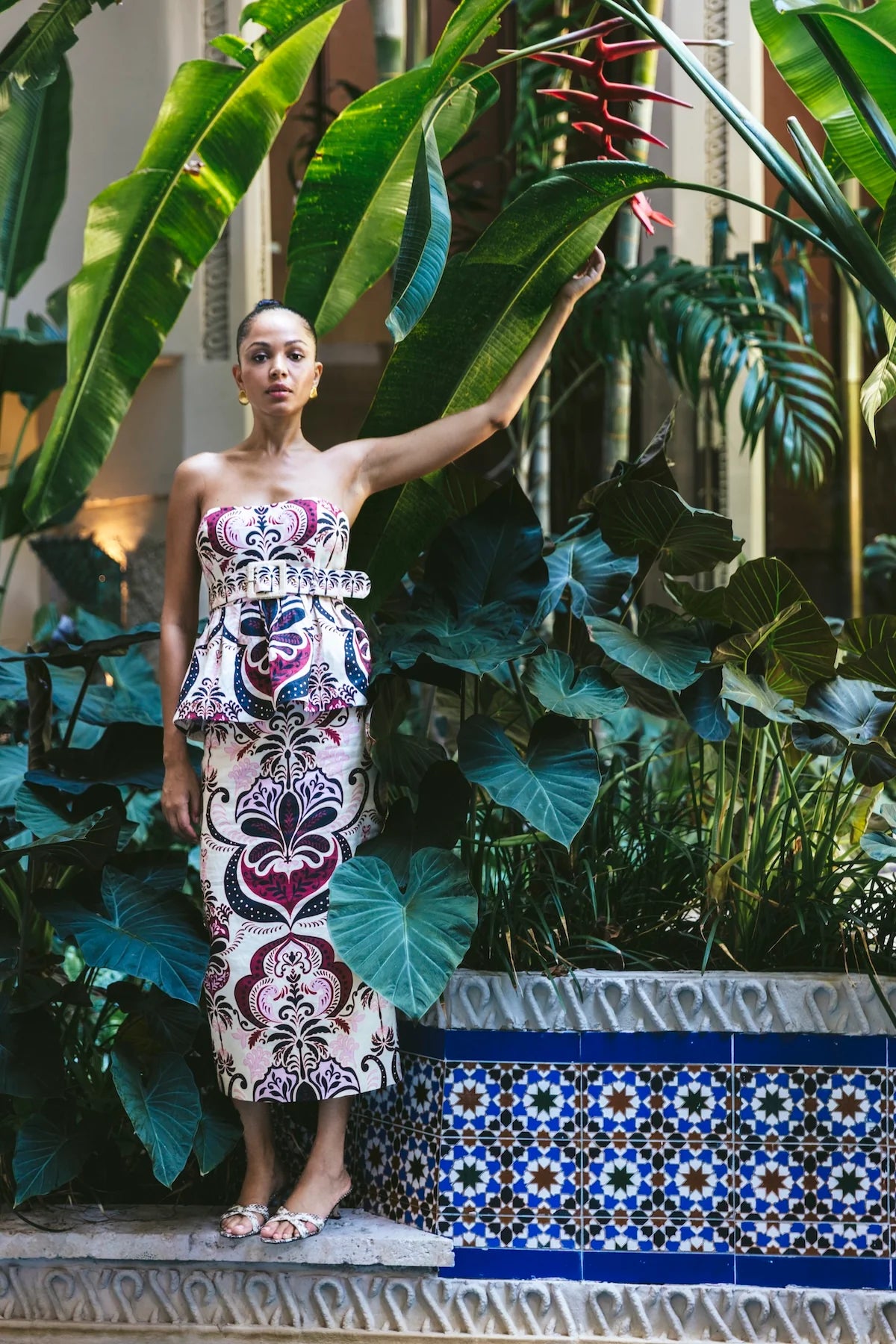 Woman in a patterned set standing in a garden with large green leaves and a tiled fountain.