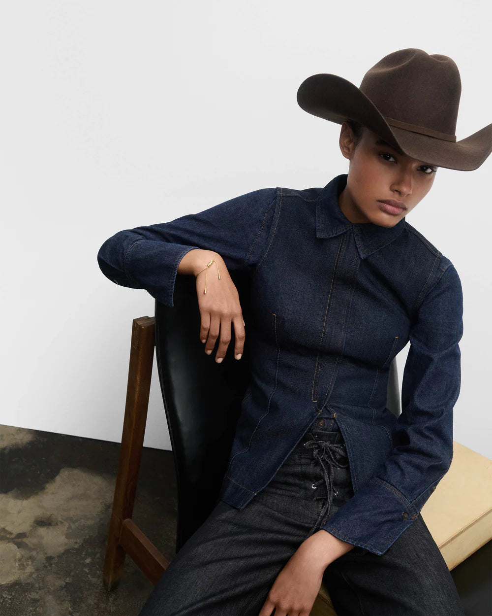 Person wearing a dark denim jacket and wide-brimmed hat sitting on a chair against a white wall.