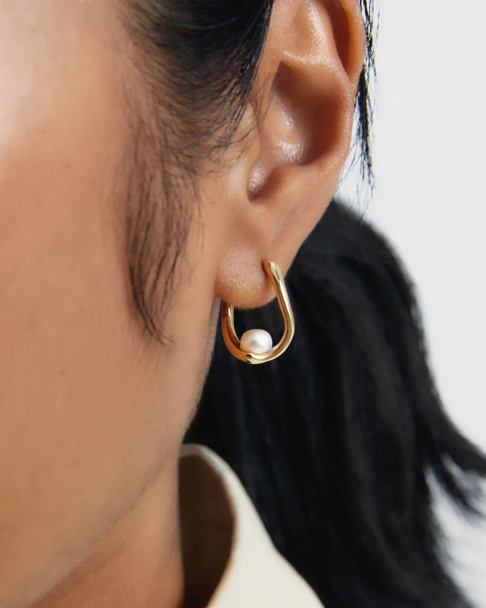 Gold hoop earring with a pearl on an ear against a neutral background