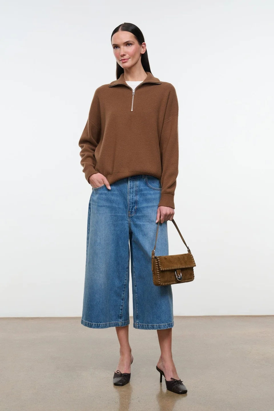 A woman stands in a studio wearing a brown quarter-zip long-sleeve sweater, blue wide-leg cropped jeans, black pointed heels, and holding a brown leather handbag.