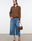 A woman stands in a studio wearing a brown quarter-zip long-sleeve sweater, blue wide-leg cropped jeans, black pointed heels, and holding a brown leather handbag.