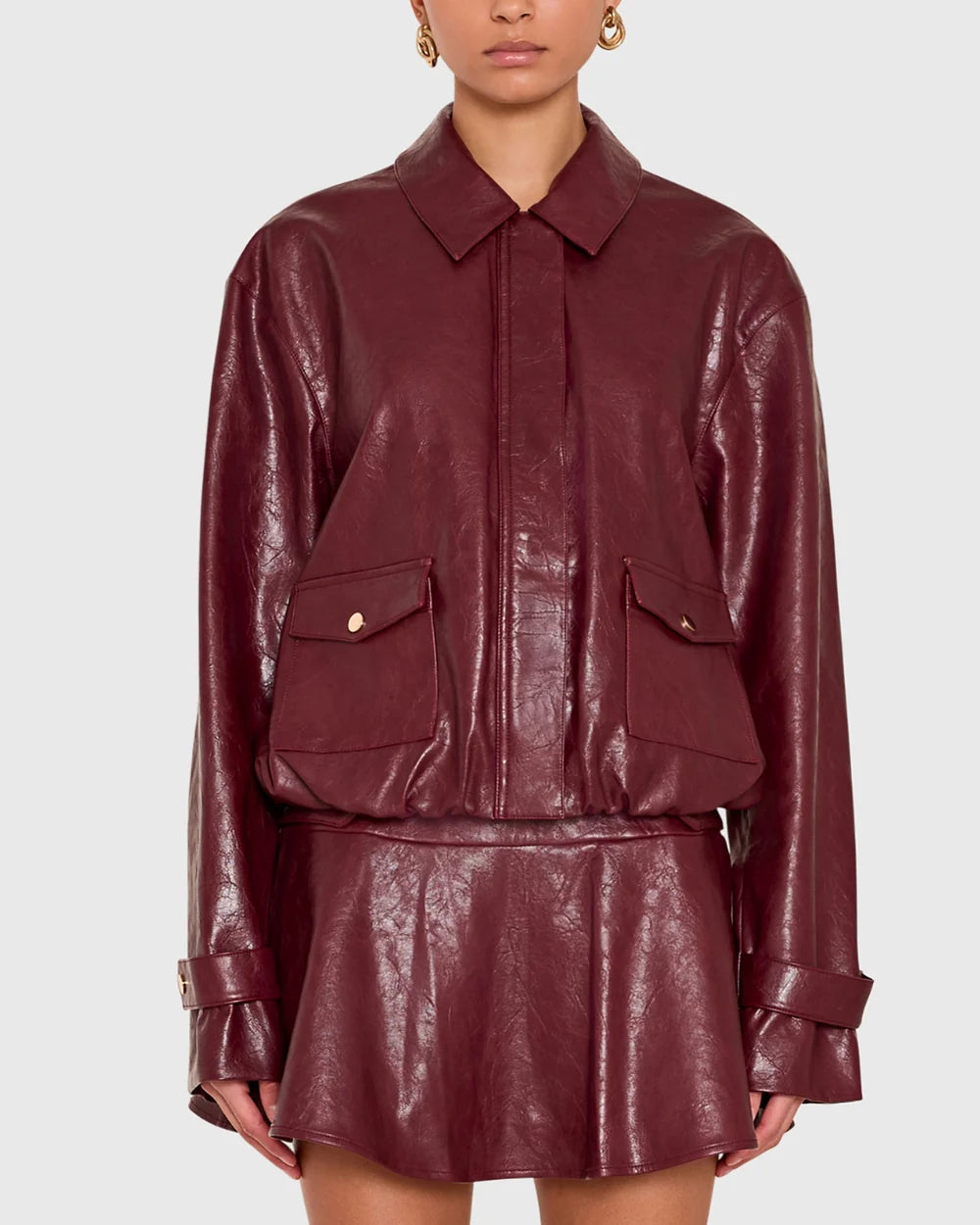 A model wearing a glossy burgundy faux-leather jacket and matching mini skirt set against a light gray studio background.
