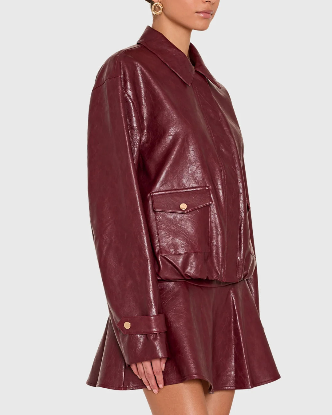 A woman wearing a glossy burgundy faux-leather jacket and matching mini skirt against a light gray studio background.