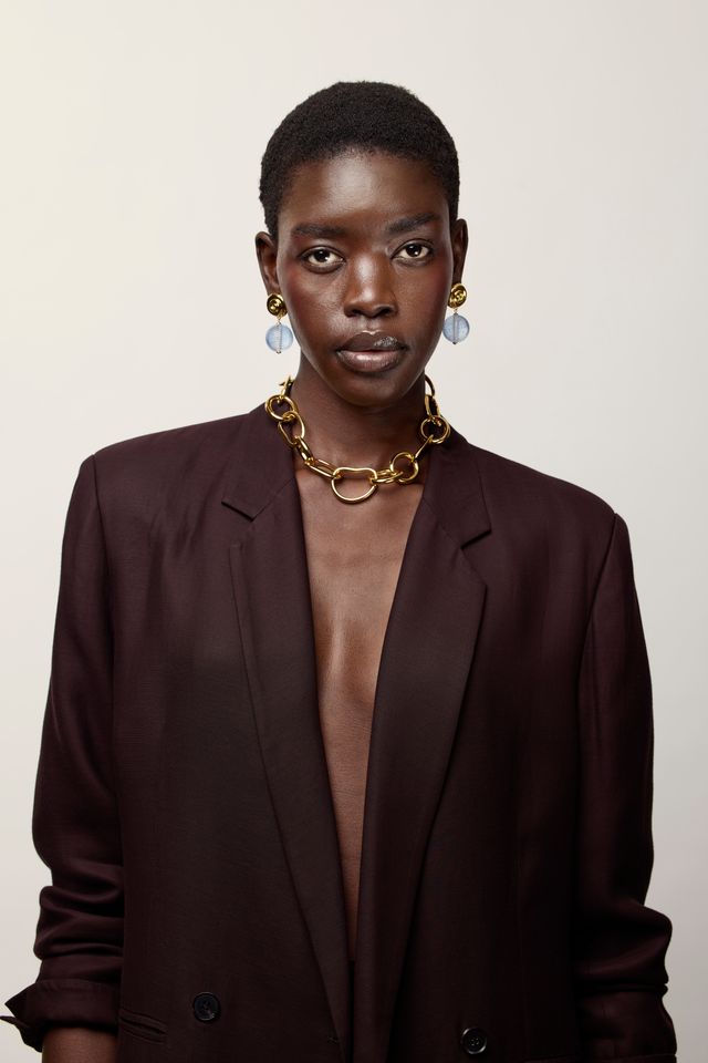 A model with short dark hair is wearing a dark brown blazer, a chunky gold chain necklace, and Luminary Earrings, posed against a solid light beige background.