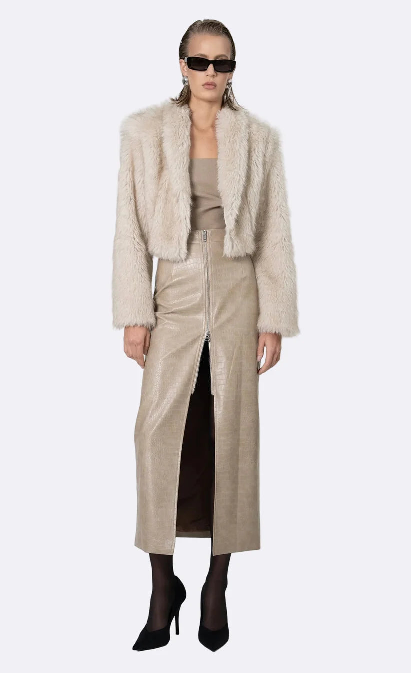 A model wearing a light beige faux fur cropped jacket over a matching beige top and high-waisted beige skirt, styled with black pointed-toe heels and sunglasses, against a light gray background.