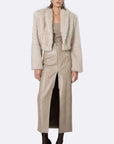 A model wearing a light beige faux fur cropped jacket over a matching beige top and high-waisted beige skirt, styled with black pointed-toe heels and sunglasses, against a light gray background.